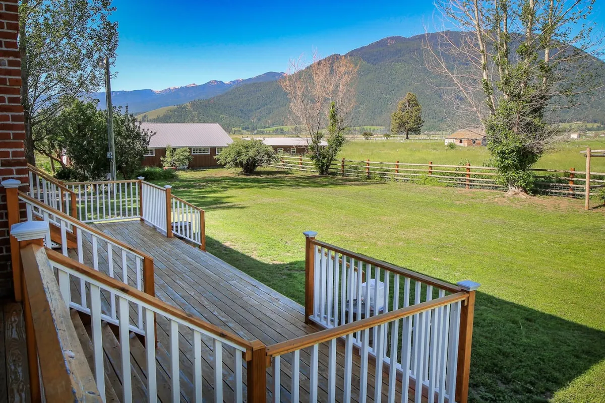 A spacious wooden deck with railings overlooks a lush, expansive lawn that stretches toward a majestic mountain backdrop. The fenced area suggests a secure, pet-friendly environment, while the serene landscape invites relaxation and outdoor enjoyment.