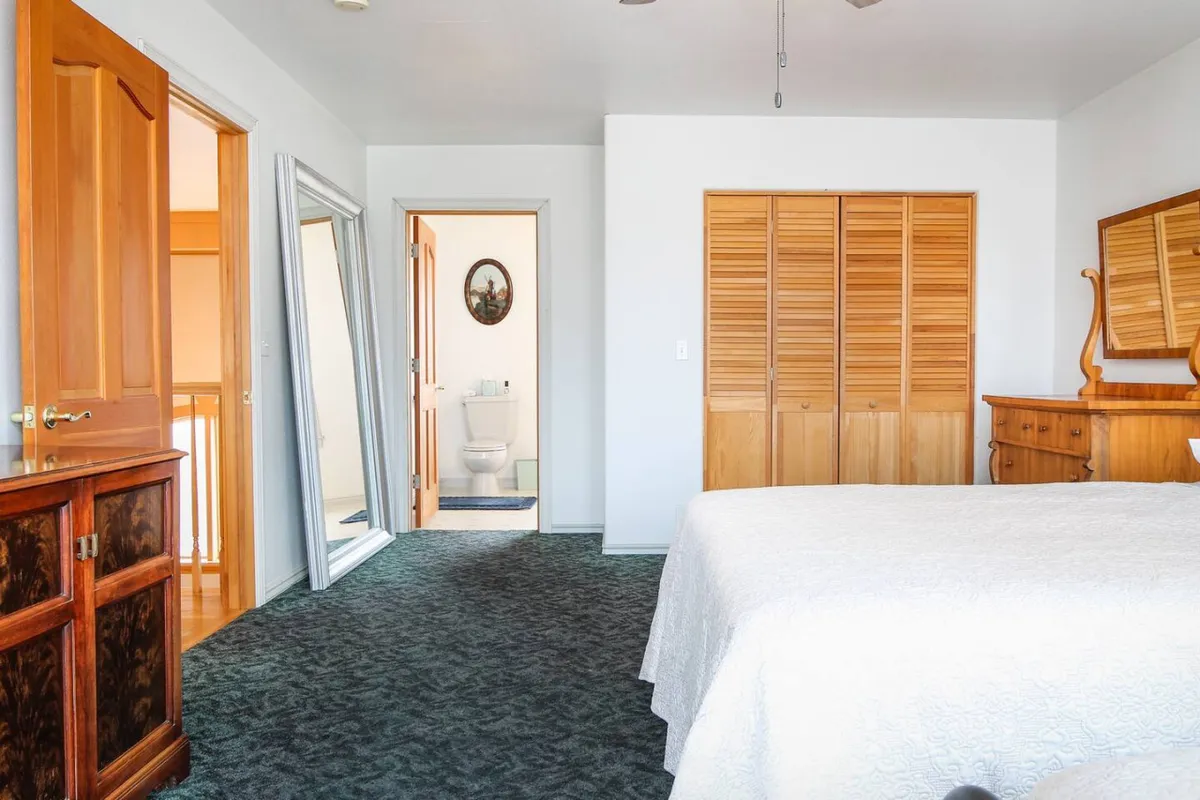 A peaceful bedroom with a bed covered in white bedding, a wooden dresser, large mirror, closet doors, and a doorway leading to an en-suite bathroom. Sunlight gently illuminates the space, enhancing the sense of calm and comfort.