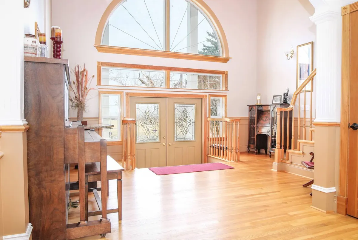 A spacious entry area with grand wooden doors, a decorative window, a piano, wooden flooring, and a sweeping staircase. The room exudes warmth and tradition, with accents that suggest music, social interaction, and family gatherings.