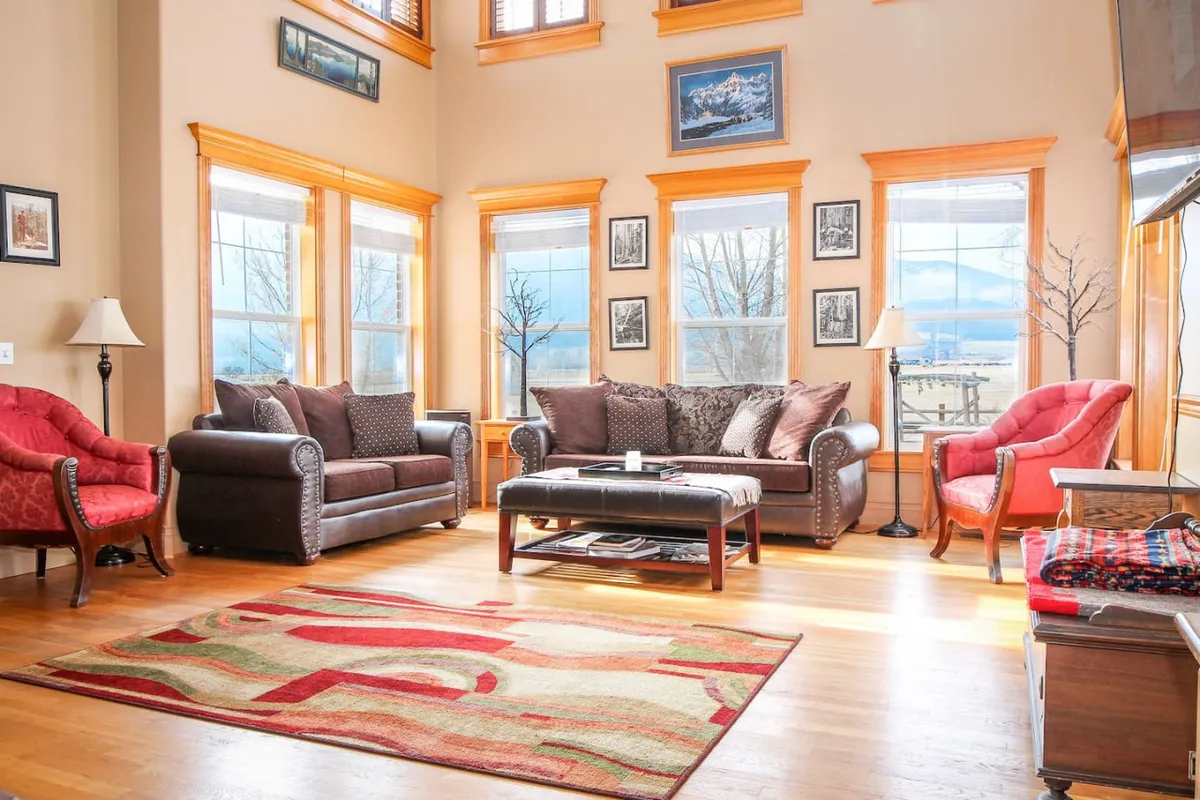 The image showcases a spacious, sunlit living area with large windows offering breathtaking views of the Elkhorn Mountains. A cozy, colorful seating arrangement invites relaxation, while vibrant area rugs and tasteful wall art add warmth and character. The scene radiates comfort and invites guests to gather, unwind, and savor the natural beauty just outside.