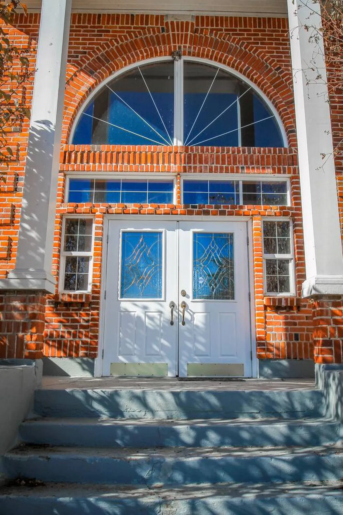 This image features the prominent double doors and arched window above the entryway of the restored brick schoolhouse, set against a charming brick facade. The exterior landscaping is inviting, and guests can be seen greeting each other, evoking a welcoming and historic ambiance.