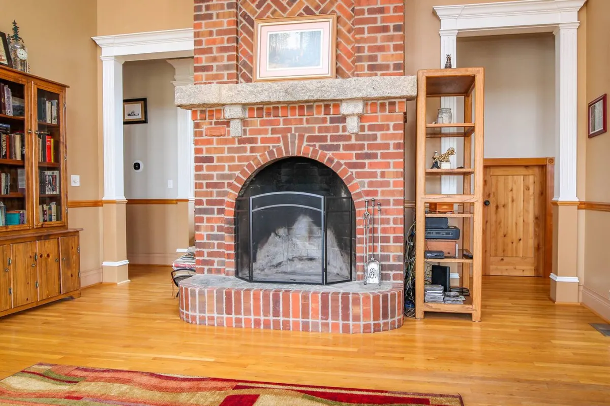 A welcoming living room featuring a prominent brick fireplace, wooden bookshelf filled with decor, and inviting seating. The scene is suffused with warm, natural light from large windows, creating a cozy space ideal for relaxation and gatherings.