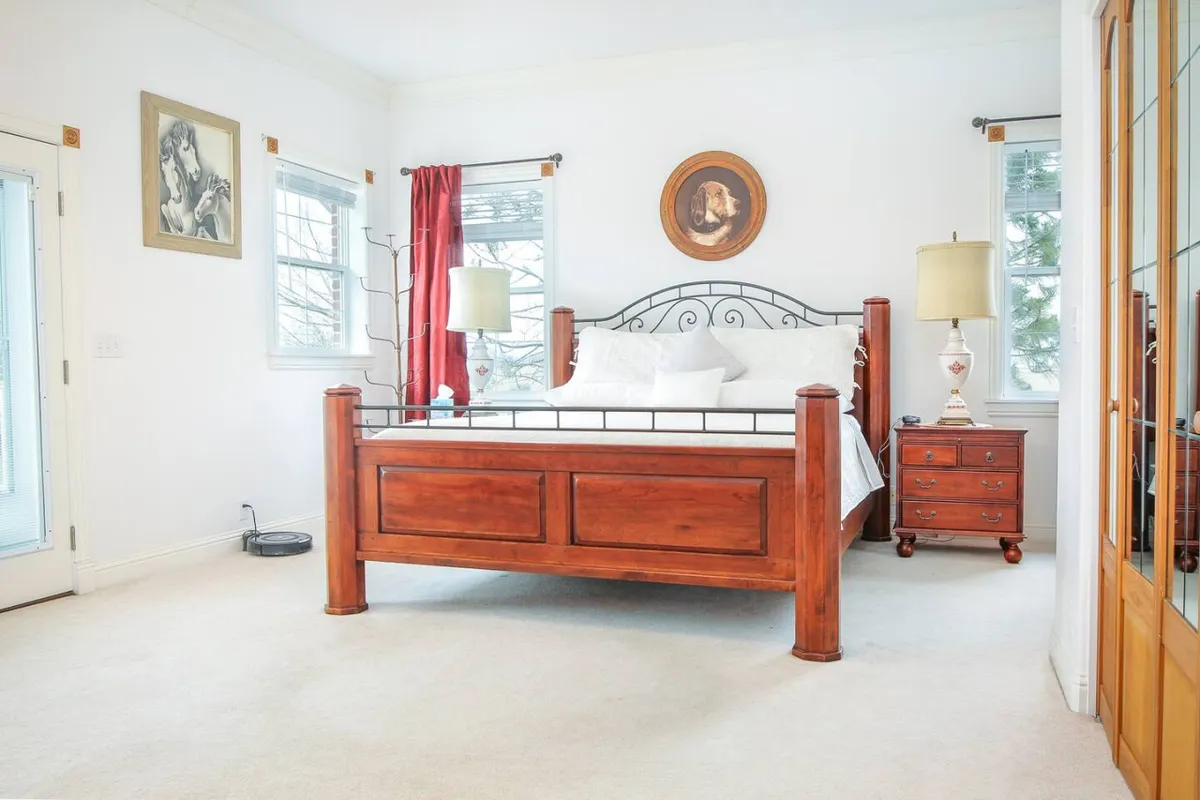 A serene bedroom featuring a classic wooden bed frame, rich red curtains, soft lighting from bedside lamps, and artwork adorning the wall. The room is bathed in subtle natural light, creating a restful and inviting atmosphere.