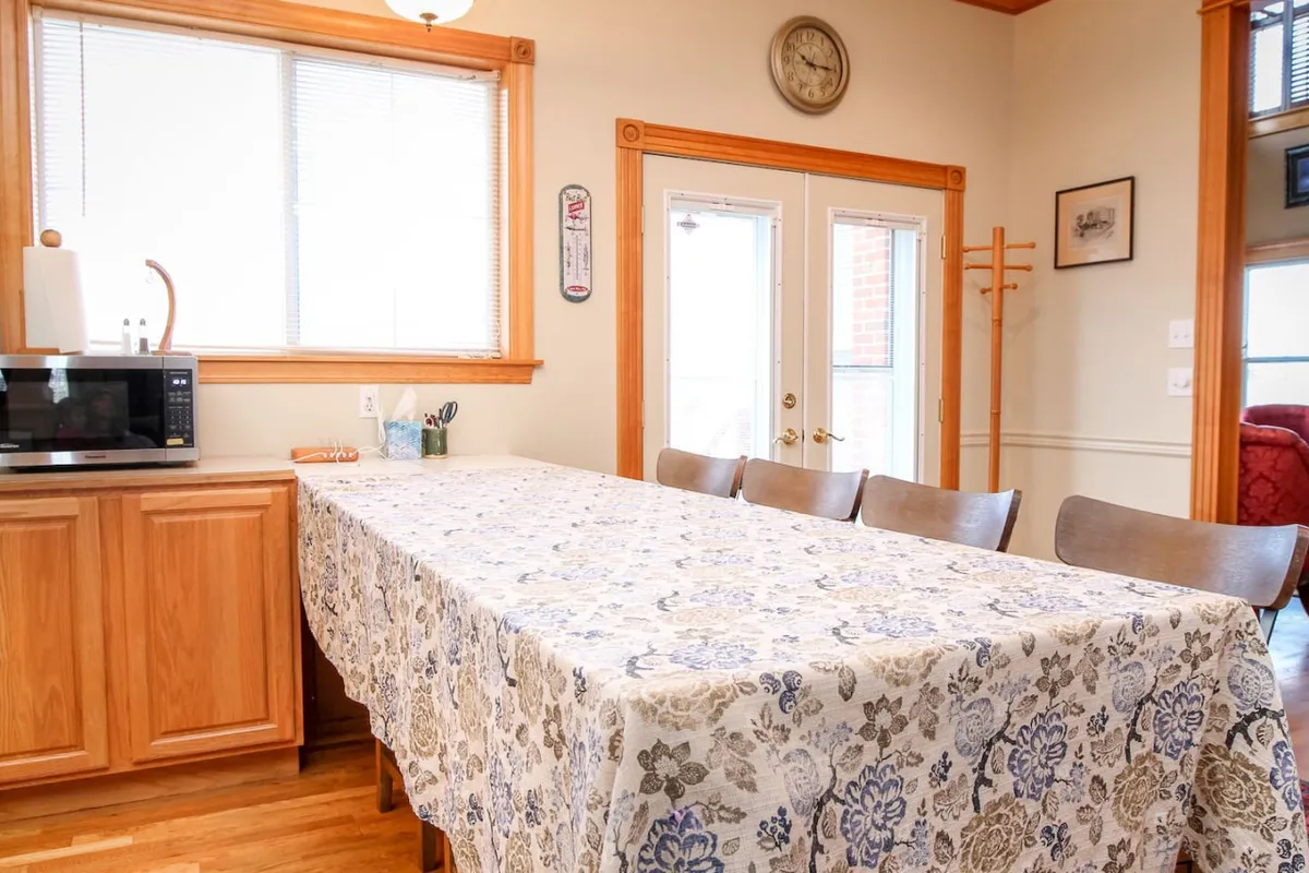 An inviting dining space with a large table, decorative tablecloth, kitchen appliances in the background, and double doors that lead outside. The room feels cozy and welcoming, ideal for casual gatherings and shared meals.