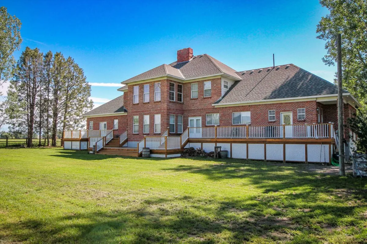 A wide view of a stately large brick house with a wrap-around porch, manicured lawn, and tall trees under expansive Oregon skies. The scene is serene and authentic, suggesting both the grandeur and the welcoming comfort of the home’s setting.