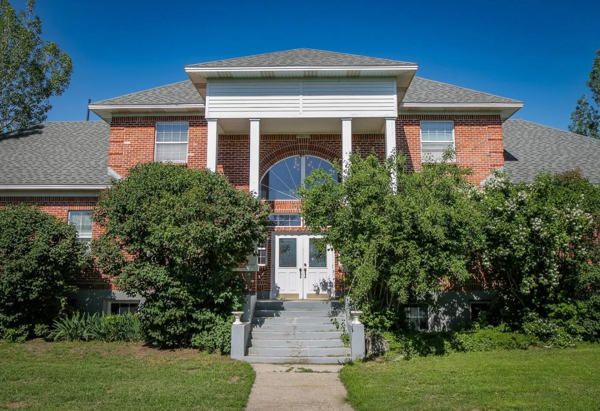 The image showcases the front entrance of the Rock Creek Schoolhouse, featuring a classic brick facade, large windows, and lush greenery framing the walkway. The welcoming porch and spacious yard are visible, suggesting ample outdoor space for both humans and pets to enjoy.