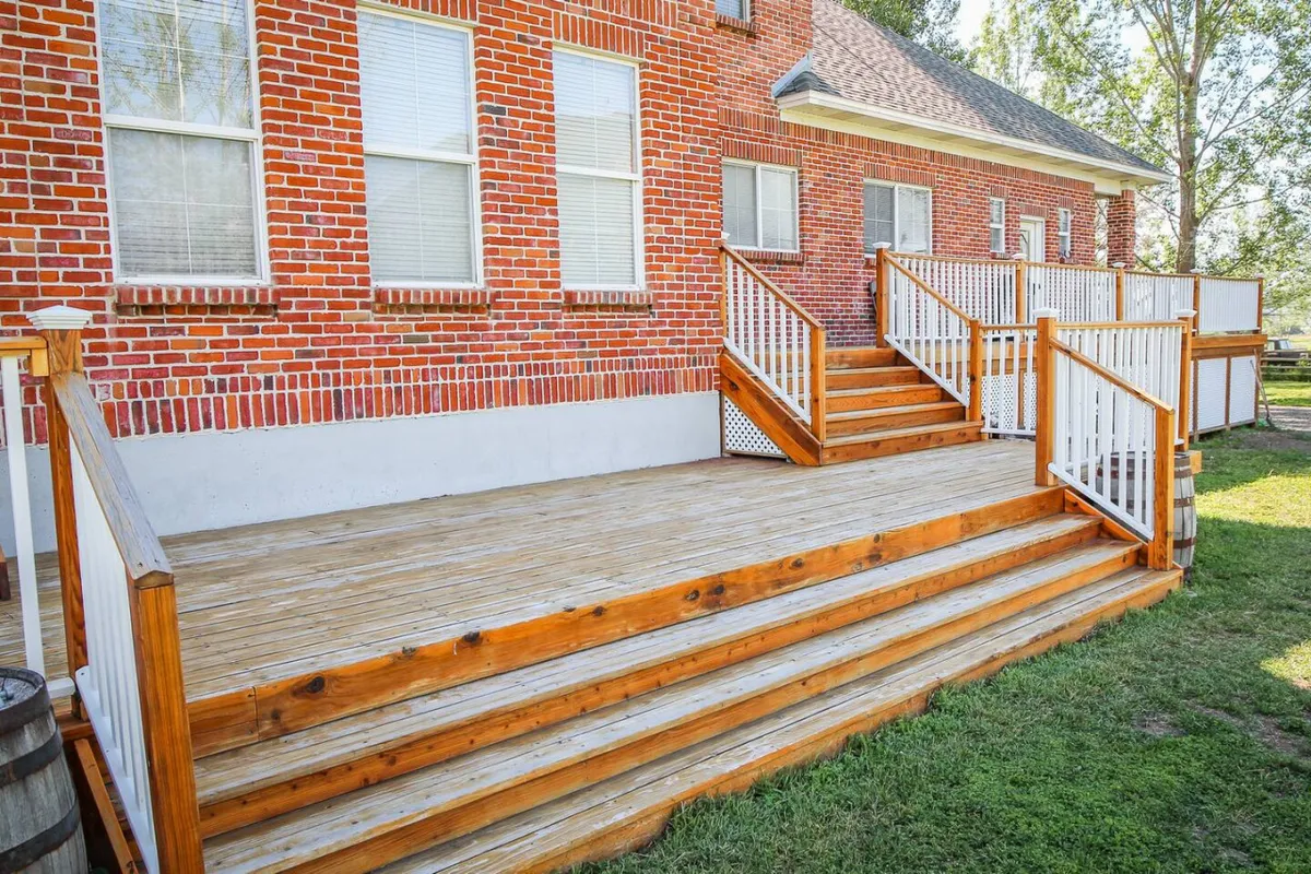 A view of the wooden back deck attached to the brick house, featuring railings and steps leading down to the grassy yard. The scene suggests a space for barbecues, group meals, and sunset relaxation, surrounded by tranquil landscaping.
