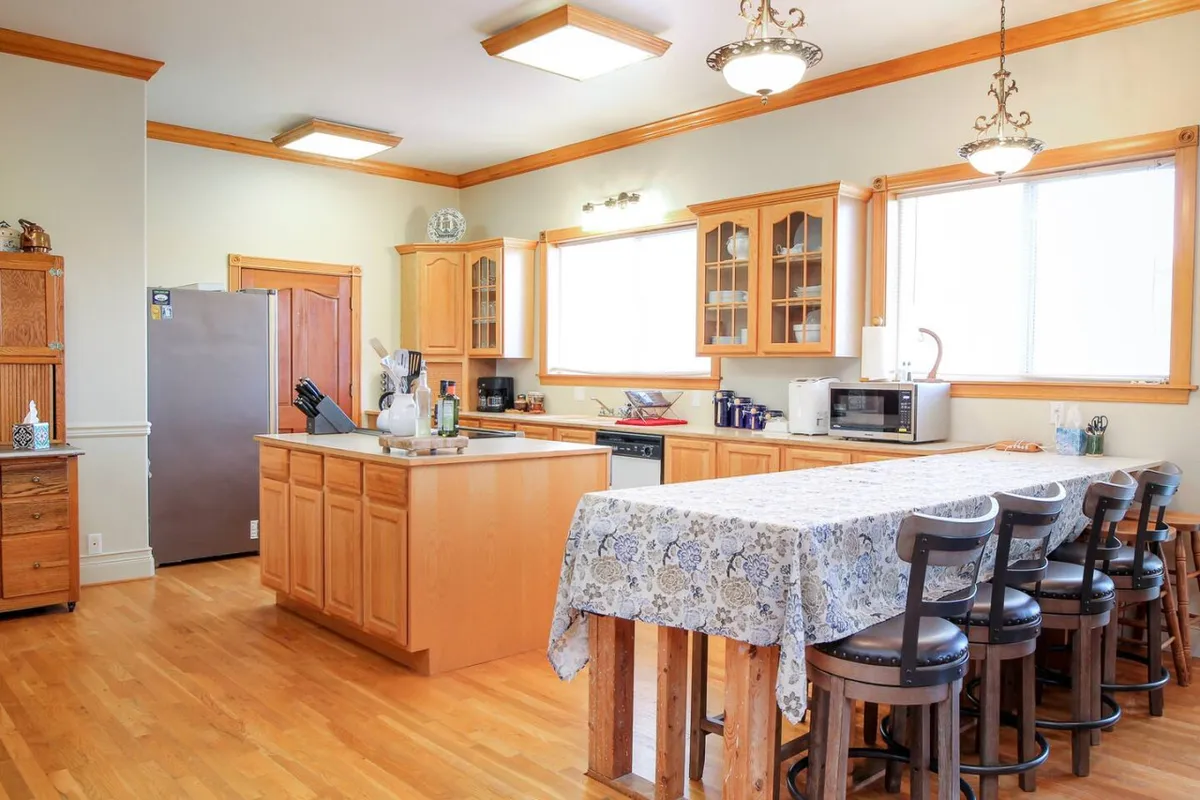 A spacious, light-filled kitchen with a large central table, well-equipped appliances, and windows letting in natural light. The space is arranged for both cooking and socializing, with plenty of room for group activities and culinary demonstrations.