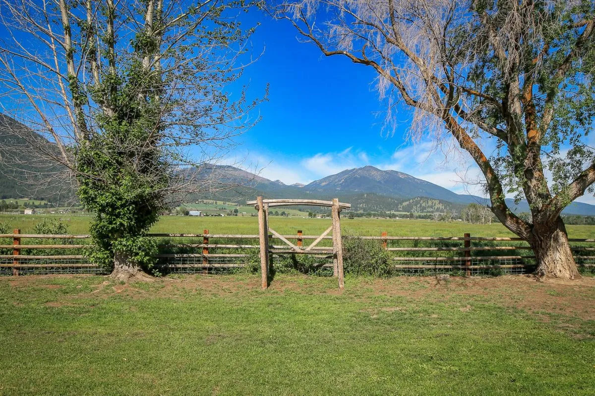 A wooden gate opens to a lush green meadow with trees and a distant mountain range, setting a tranquil yet adventurous tone. The scene suggests access to miles of hiking trails and natural beauty in the Elkhorn Mountains.