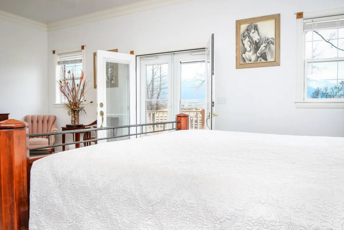 A cozy bedroom with a bed dressed in a white quilt, an inviting armchair beside it, and tasteful artwork on the wall. Sunlight streams through open doors that lead outside, offering views and a sense of connection with nature.