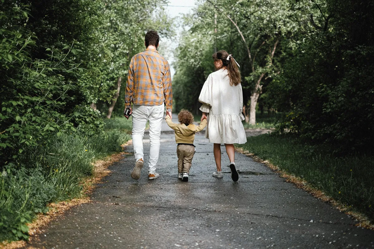 A family, including children and adults, walks together down a tree-lined pathway, surrounded by lush green trees. The mood is happy and connected, emphasizing time spent outdoors bonding and exploring nature as a group.